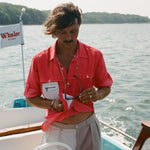 Man on a boat wearing a red button-up shirt with a white mesh pocket and holding sunglasses. A white flag with red text reading 'Whaler by Tombolo' is visible in the background.