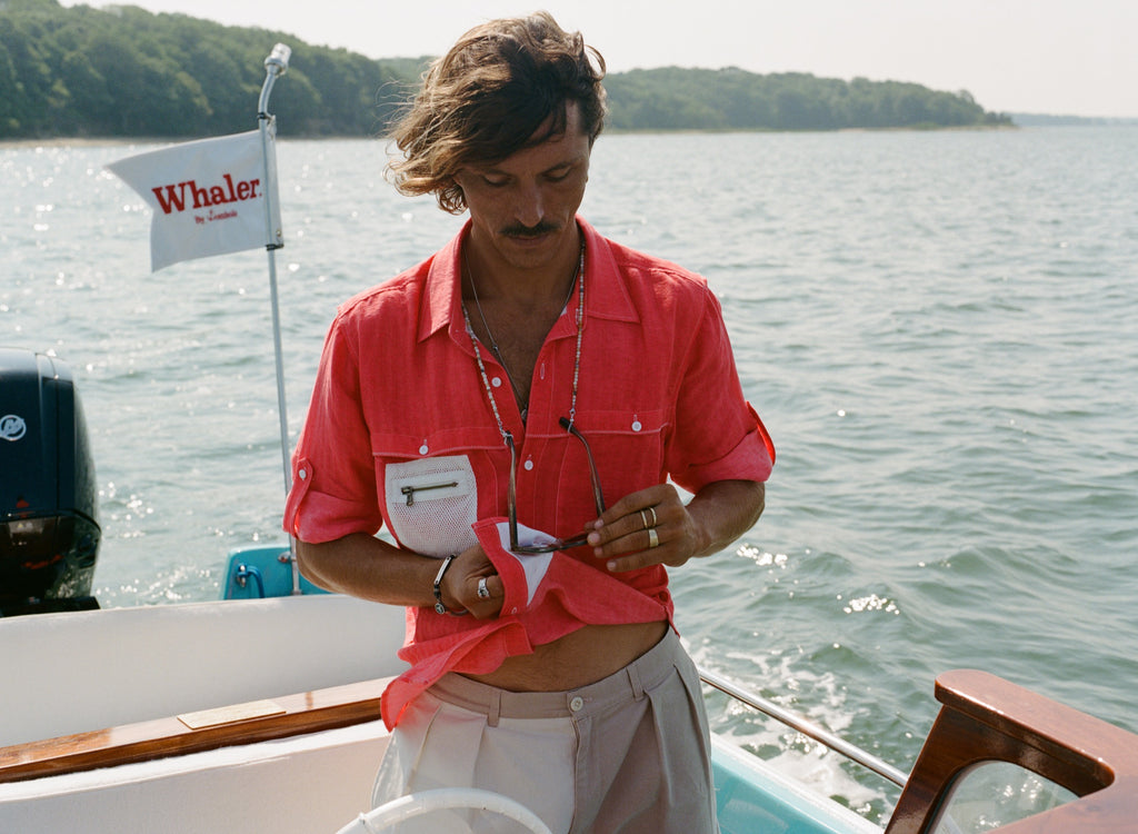 Man on a boat wearing a red button-up shirt with a white mesh pocket and holding sunglasses. A white flag with red text reading 'Whaler by Tombolo' is visible in the background.