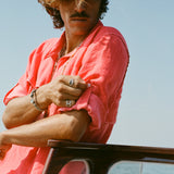 Man rolling up the sleeve of a red button-up shirt while leaning on a wooden boat railing.