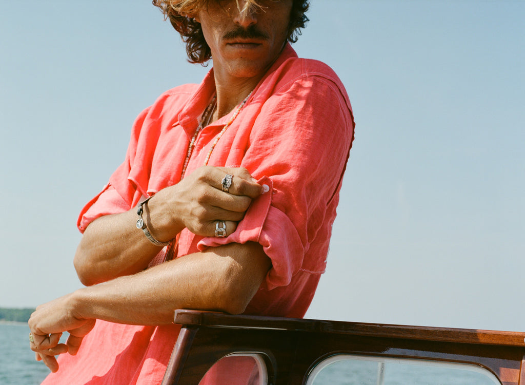 Man rolling up the sleeve of a red button-up shirt while leaning on a wooden boat railing.