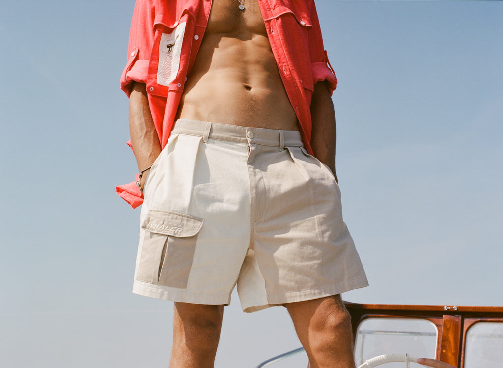 Man wearing two-tone pleated shorts in khaki and off-white, featuring a flap pocket on the left with white 'Boston Whaler' embroidery. He has a red button-down shirt open on top.
