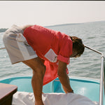Man on a boat bending forward while adjusting a white cover. He wears two-tone khaki and off-white shorts with a side flap pocket and a red fishing shirt with a white mesh pocket.