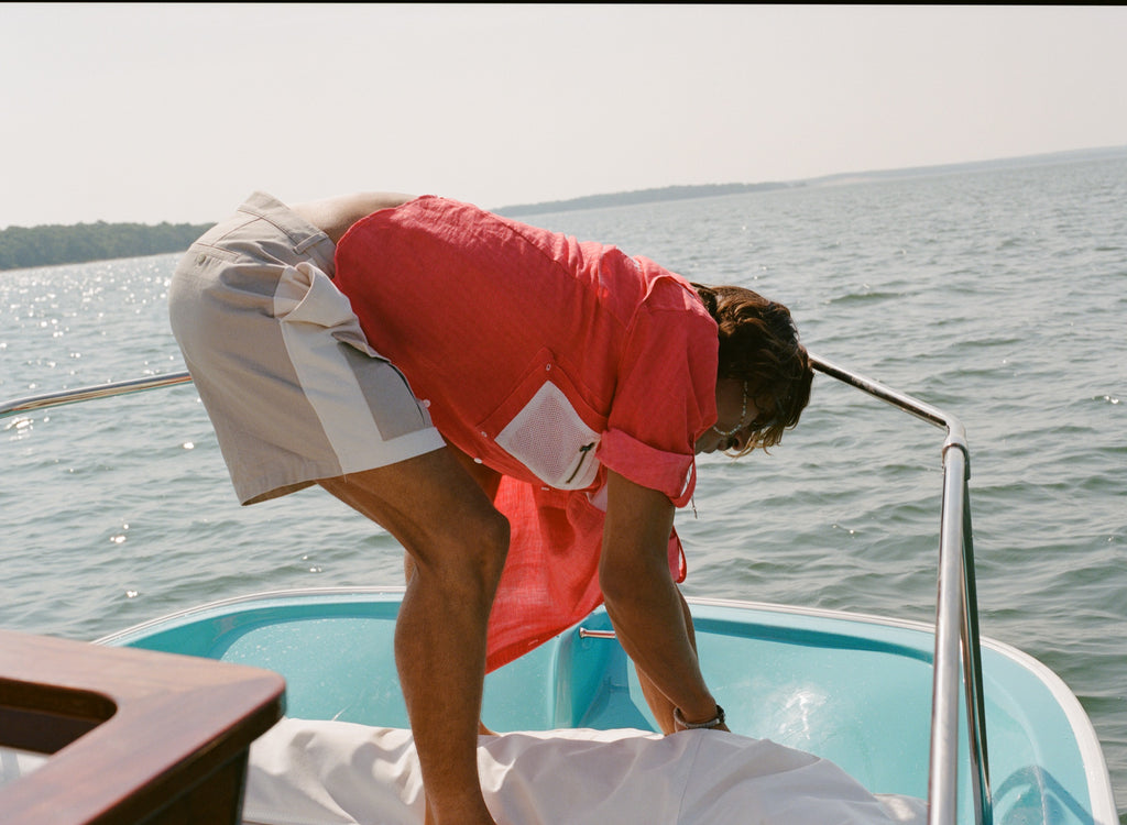 Man on a boat bending forward while adjusting a white cover. He wears two-tone khaki and off-white shorts with a side flap pocket and a red fishing shirt with a white mesh pocket.