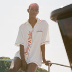 Woman sitting on the edge of a boat wearing a white terry short-sleeve zip up cabana with red 'Boston Whaler' text and red contrast stitching.