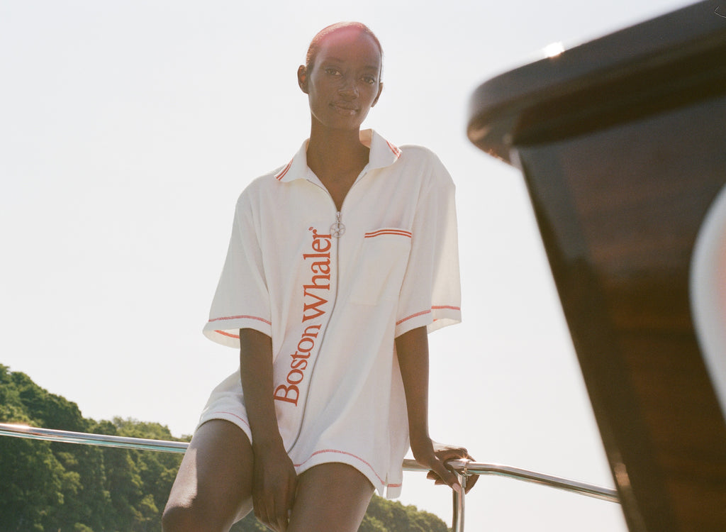 Woman sitting on the edge of a boat wearing a white terry short-sleeve zip up cabana with red 'Boston Whaler' text and red contrast stitching.