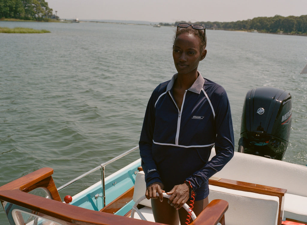 Woman wearing a navy sun shirt with white piping and a striped collar, standing at the helm of a boat.