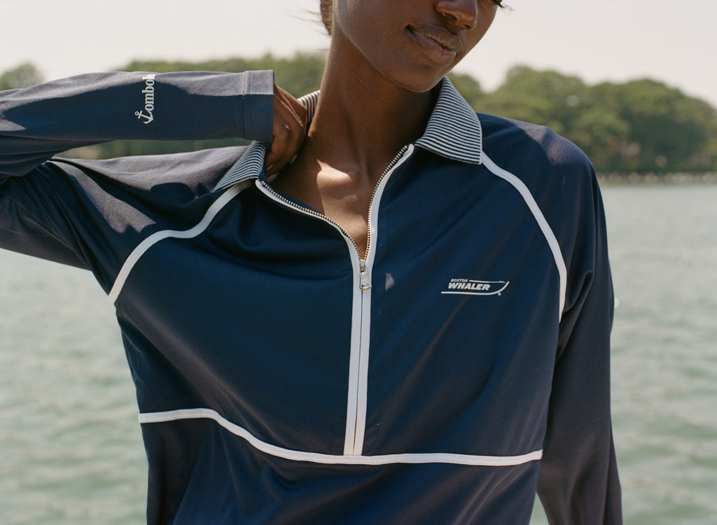 Close-up of a woman wearing a navy long-sleeve sun shirt with white piping and a striped collar. White embroidered text on the left chest reads 'Boston Whaler,' and 'Tombolo' is stitched on the sleeve.