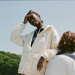 A woman wears a cream hooded rain jacket with tan trim and front snap pockets layered over a navy top, standing on a boat. A man with a two-tone polo sits in front of her.