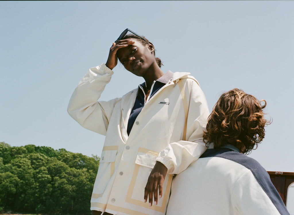 A woman wears a cream hooded rain jacket with tan trim and front snap pockets layered over a navy top, standing on a boat. A man with a two-tone polo sits in front of her.