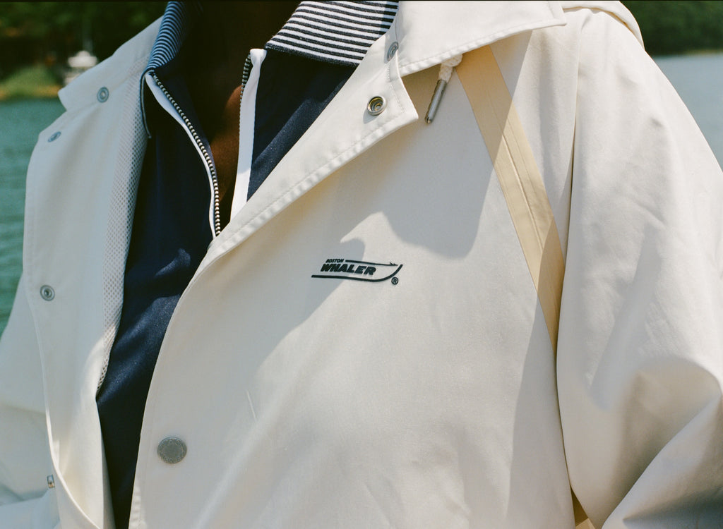 Close-up of a cream rain jacket with tan trim and Boston Whaler logo, worn over a navy top with a striped collar and white piping.