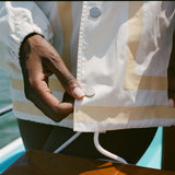 Close-up of the hem of a cream jacket with tan trim and branded snap buttons reading Boston Whaler. A white drawcord hangs below, and a woman's hand is shown adjusting the bottom snap.