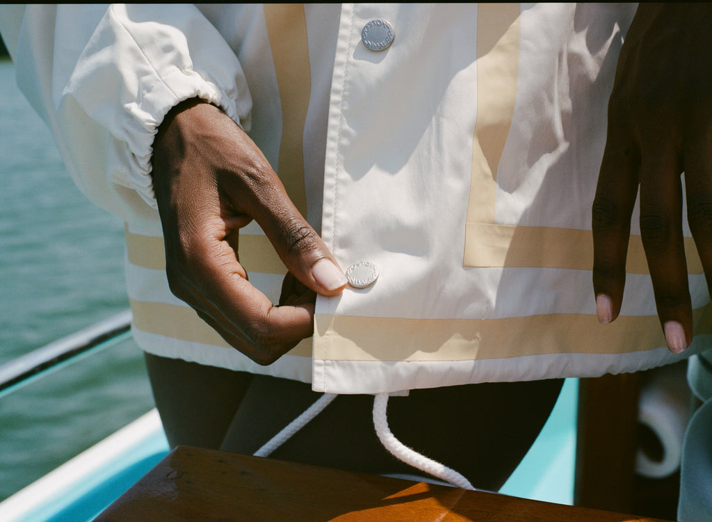Close-up of the hem of a cream jacket with tan trim and branded snap buttons reading Boston Whaler. A white drawcord hangs below, and a woman's hand is shown adjusting the bottom snap.