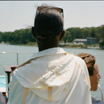 Woman facing forward at the helm of a boat, wearing a cream hooded jacket with tan trim and mesh lining.