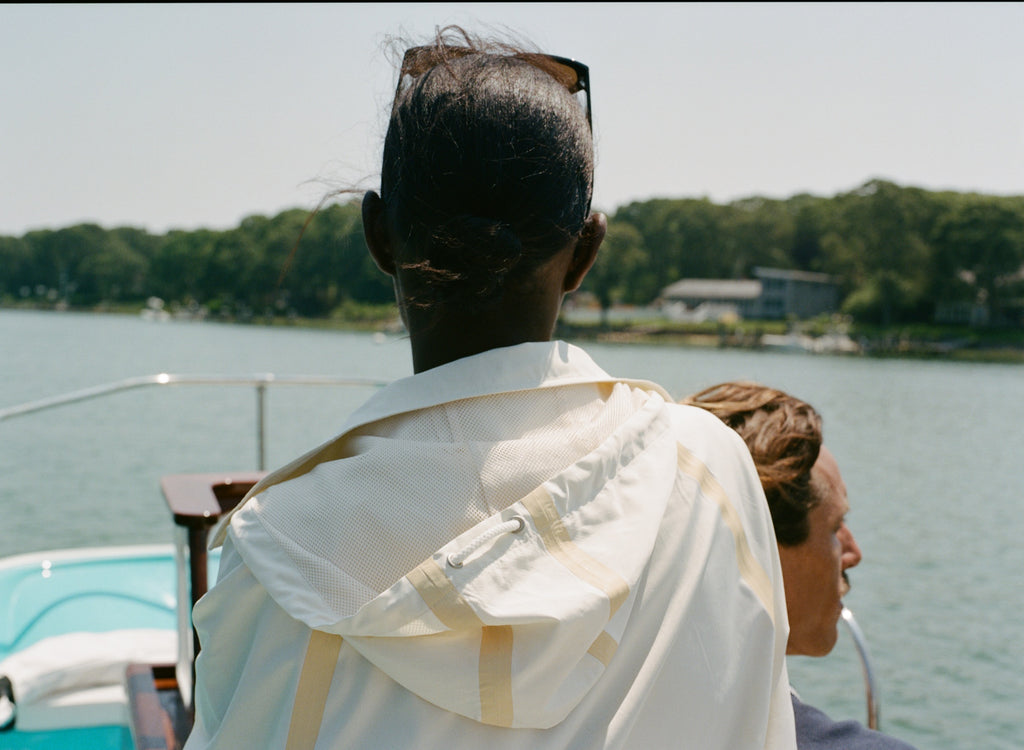 Woman facing forward at the helm of a boat, wearing a cream hooded jacket with tan trim and mesh lining.