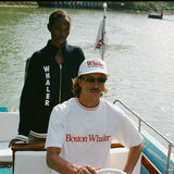 Man at the wheel of a boat wearing a white t-shirt with 'Boston Whaler' embroidered in red across the chest and a matching red-and-white knit collar. Woman behind him wears a navy zip-up sweater with large vertical 'WHALER' text.