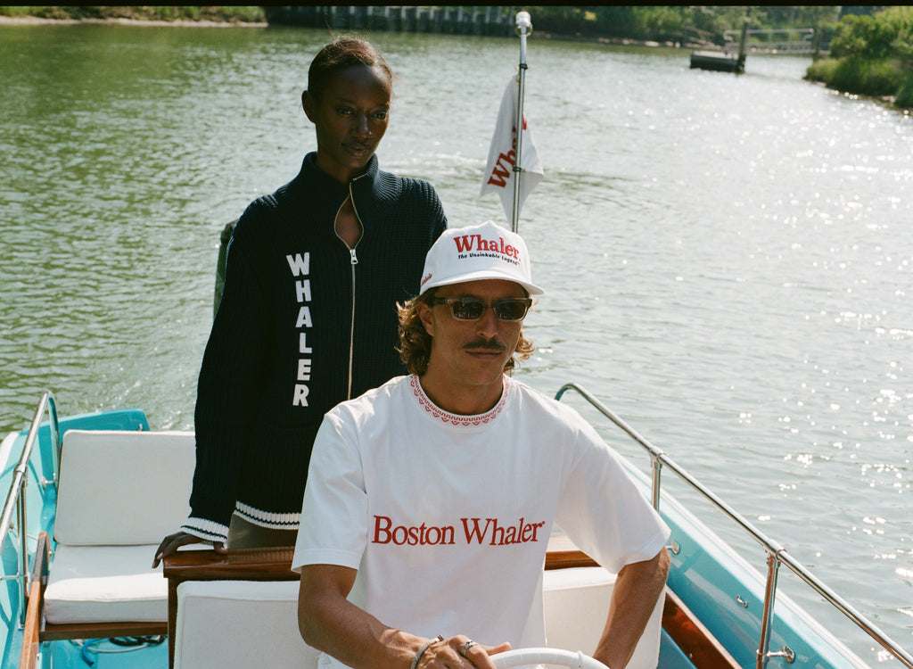 Man at the wheel of a boat wearing a white t-shirt with 'Boston Whaler' embroidered in red across the chest and a matching red-and-white knit collar. Woman behind him wears a navy zip-up sweater with large vertical 'WHALER' text.