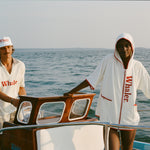 Man wearing a white five-panel “Whaler” cap and matching white polo shirt stands at the helm of a boat. Next to him, a woman leans against the cabin wearing a white hooded terry zip-up with red “Whaler” text down the front and red trim.