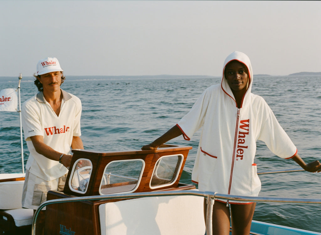 Man wearing a white five-panel 'Whaler' cap and matching white polo shirt stands at the helm of a boat. Next to him, a woman leans against the cabin wearing a white hooded terry zip-up with red 'Whaler' text down the front and red trim. A white flag with red 'Whaler' branding waves in the background.