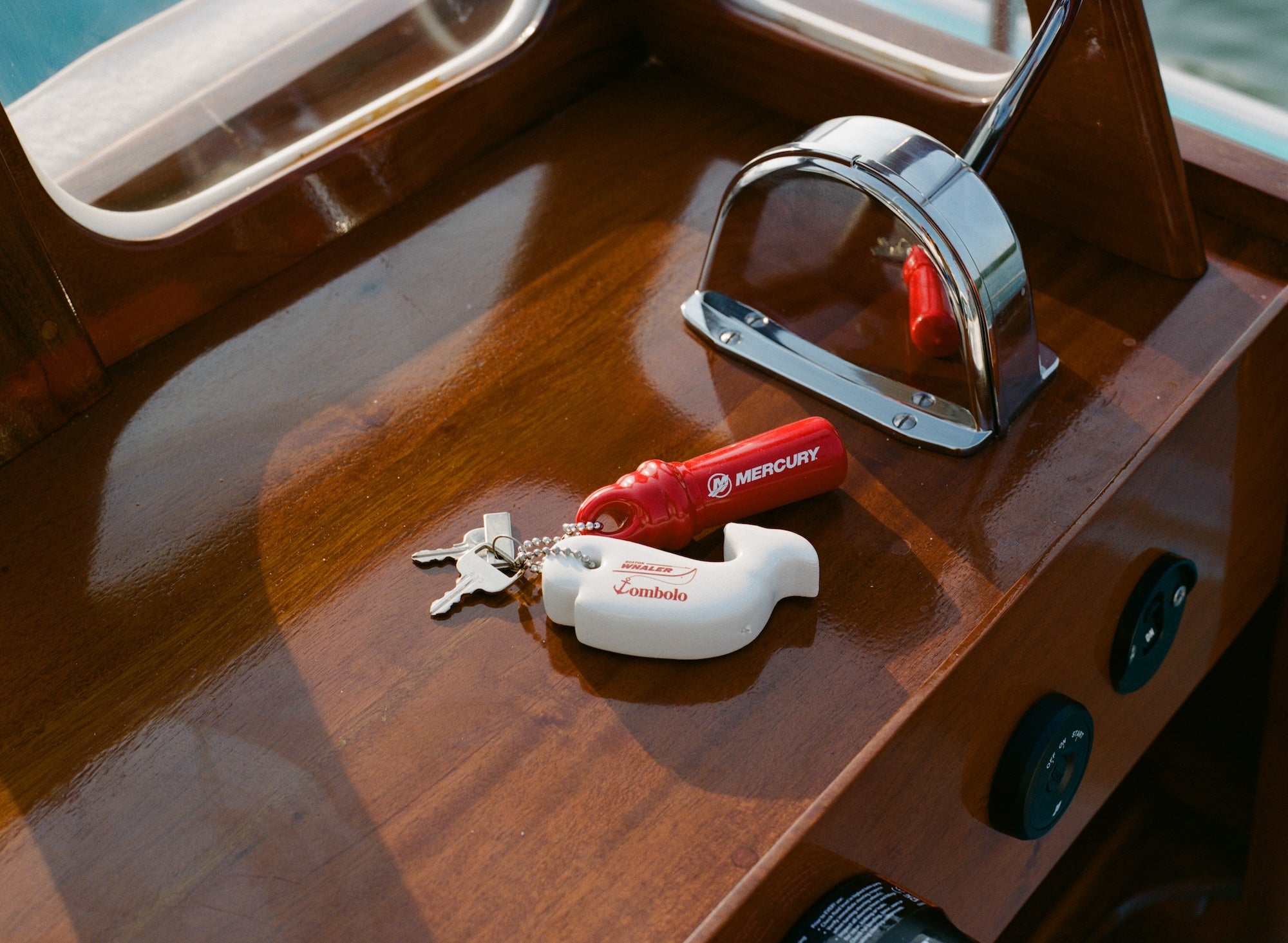 Close-up of boat dashboard with a whale-shaped white key float labeled 'Boston Whaler' and 'Tombolo' attached to a keyring. Also attached is a red Mercury engine kill switch.