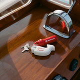 Close-up of boat dashboard with a whale-shaped white key float labeled 'Boston Whaler' and 'Tombolo' attached to a keyring. Also attached is a red Mercury engine kill switch.