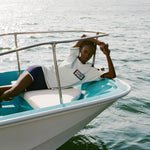 Woman laying at edge of a boat wearing the light blue cotton mesh shirt with navy shoulder panels and a 'Boston Whaler' logo on the chest, paired with matching navy shorts with white trim. 