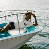 Woman laying at edge of a boat wearing the light blue cotton mesh shirt with navy shoulder panels and a 'Boston Whaler' logo on the chest, paired with matching navy shorts with white trim. 