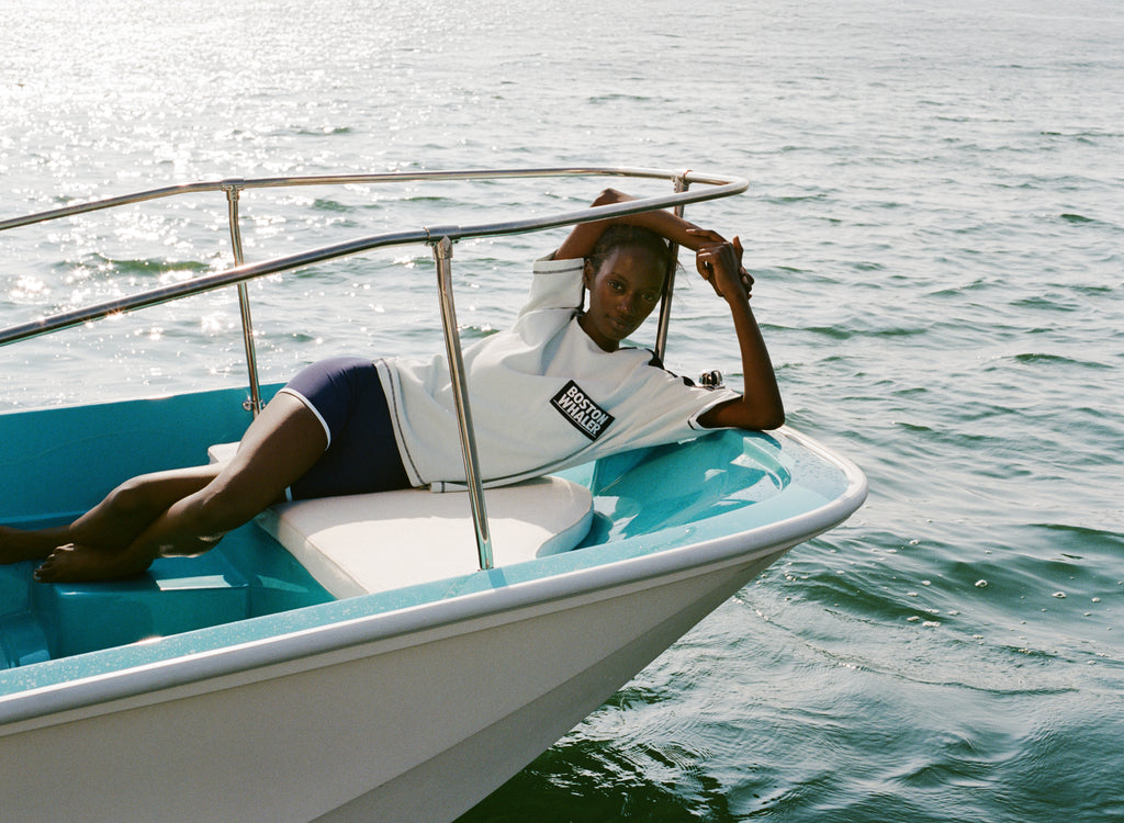 Woman laying at edge of a boat wearing the light blue cotton mesh shirt with navy shoulder panels and a 'Boston Whaler' logo on the chest, paired with matching navy shorts with white trim. 
