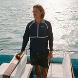 A man stands on a boat wearing a navy long-sleeve sun shirt with white piping, a striped collar, and a quarter zip. White embroidered text on the left chest reads 'Whaler'; 'Tombolo' is embroidered on the right sleeve and 'UPF 50+' on the left sleeve.