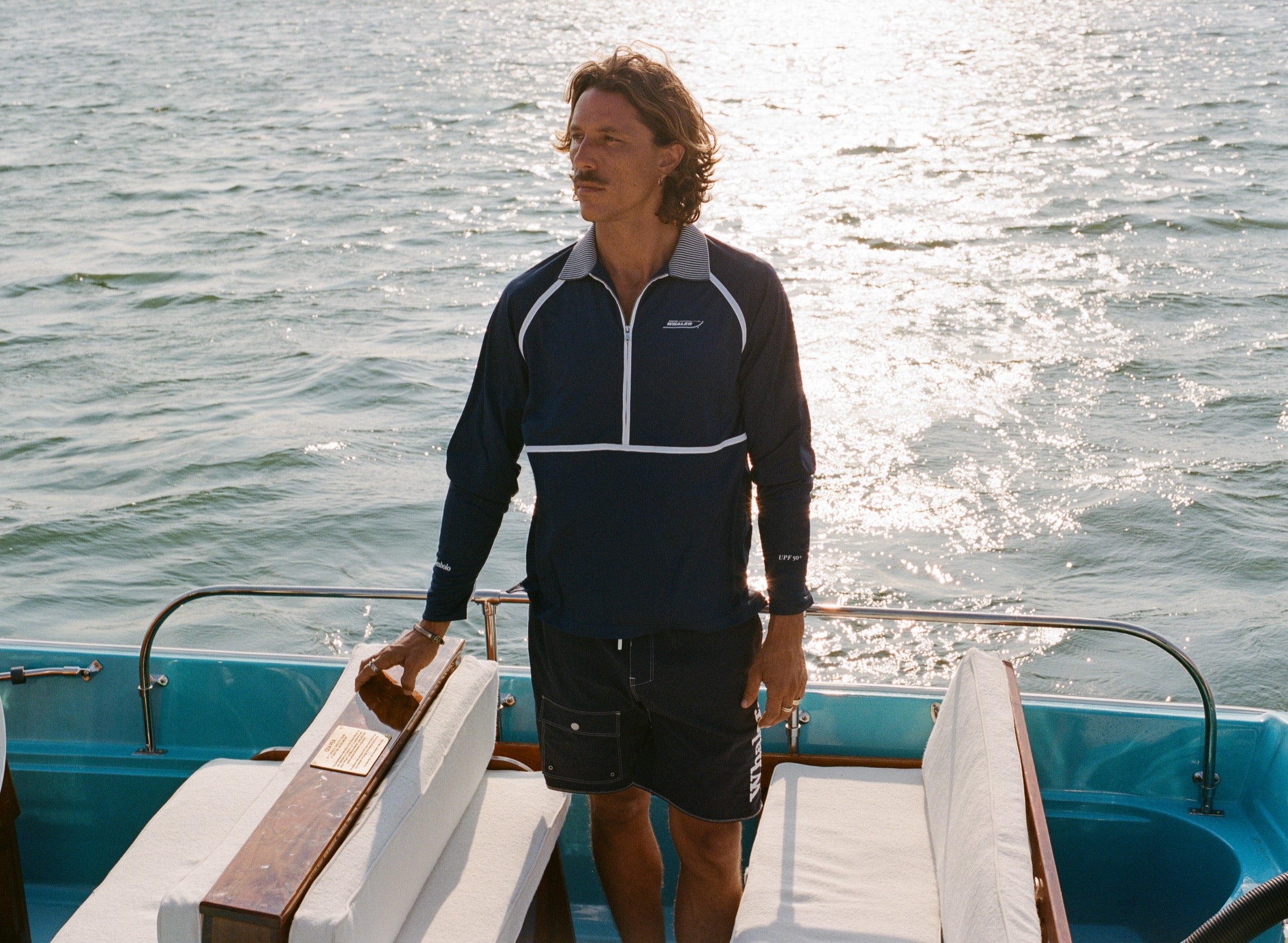 A man stands on a boat wearing a navy long-sleeve sun shirt with white piping, a striped collar, and a quarter zip. White embroidered text on the left chest reads 'Whaler'; 'Tombolo' is embroidered on the right sleeve and 'UPF 50+' on the left sleeve.