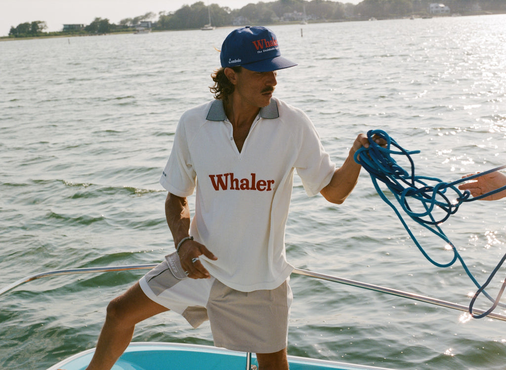 Man standing on a boat holding a coiled blue rope, wearing a blue 'Whaler' cap, a white 'Whaler' shirt, and beige color block shorts. 