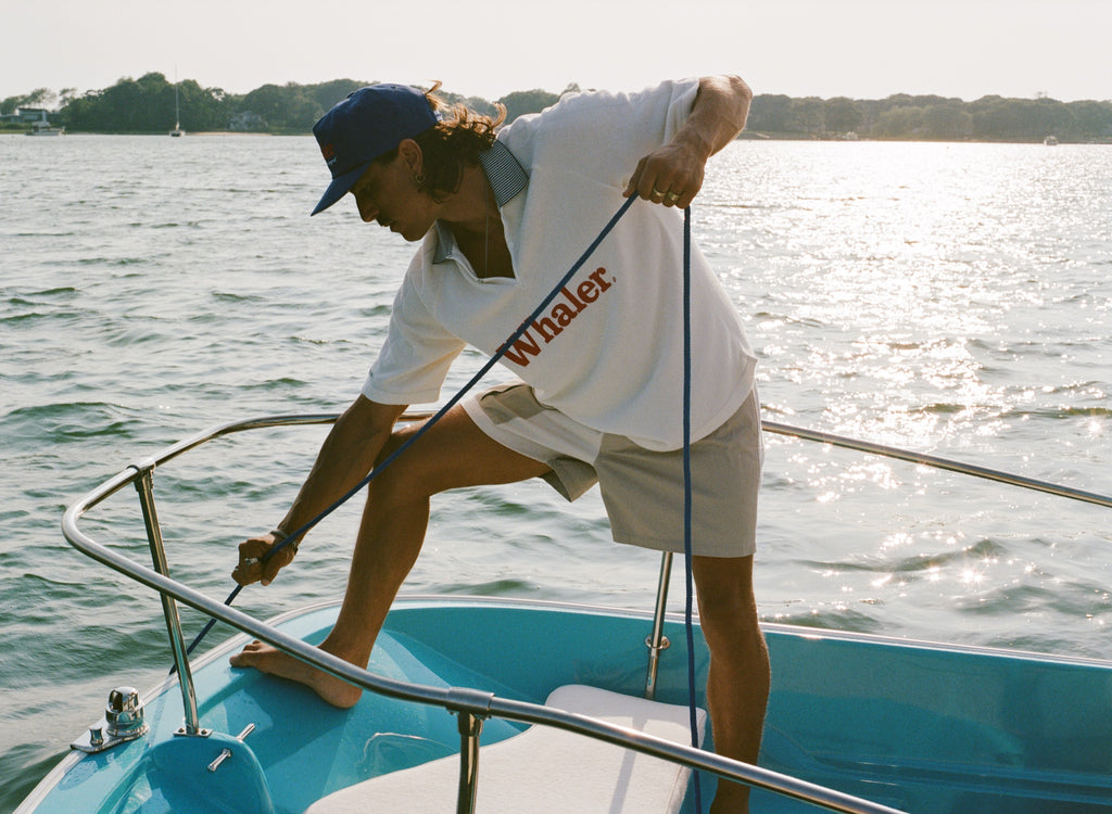 Man on a boat pulling a rope while wearing a blue 'Whaler' hat, white 'Whaler' polo, and beige shorts.