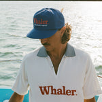 Man on a boat wearing a blue 'Whaler' hat and a white collared shirt with 'Whaler' printed on it.