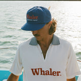 Man on a boat wearing a blue 'Whaler' hat and a white collared shirt with 'Whaler' printed on it.
