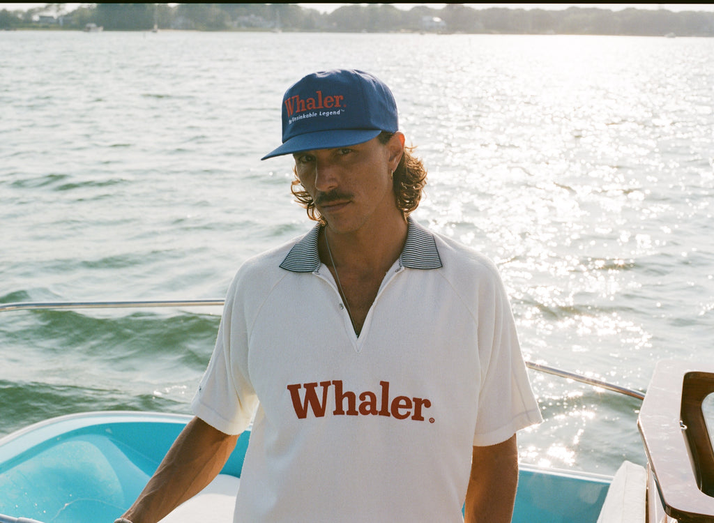 Man standing on a boat wearing a blue 'Whaler' cap and a white collared 'Whaler' polo.