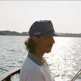 Man in a blue 'Whaler' cap and white collared shirt standing on a boat. The side of the cap reads 'Tombolo' in white embroidery.