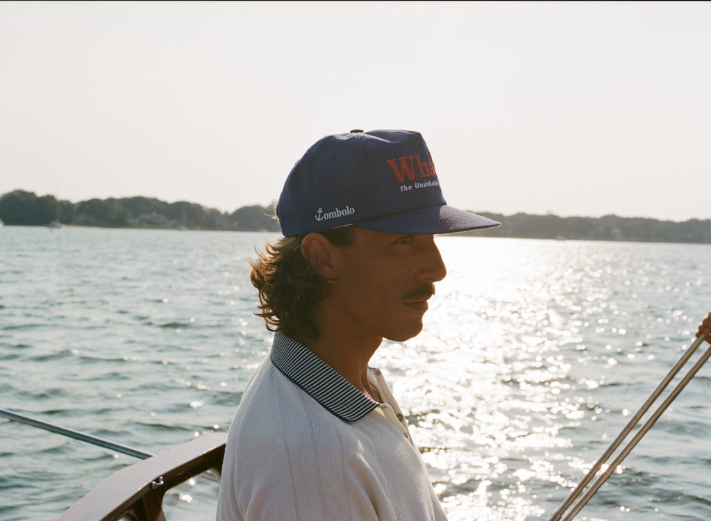 Man in a blue 'Whaler' cap and white collared shirt standing on a boat. The side of the cap reads 'Tombolo' in white embroidery.