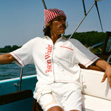 Man wearing white short-sleeve terry cloth zip-up shirt with red vertical 'Boston Whaler' text and red contrast stitching, while sitting on a boat. He also wears a red-and-white striped beanie with a 'Whaler' logo across the front.