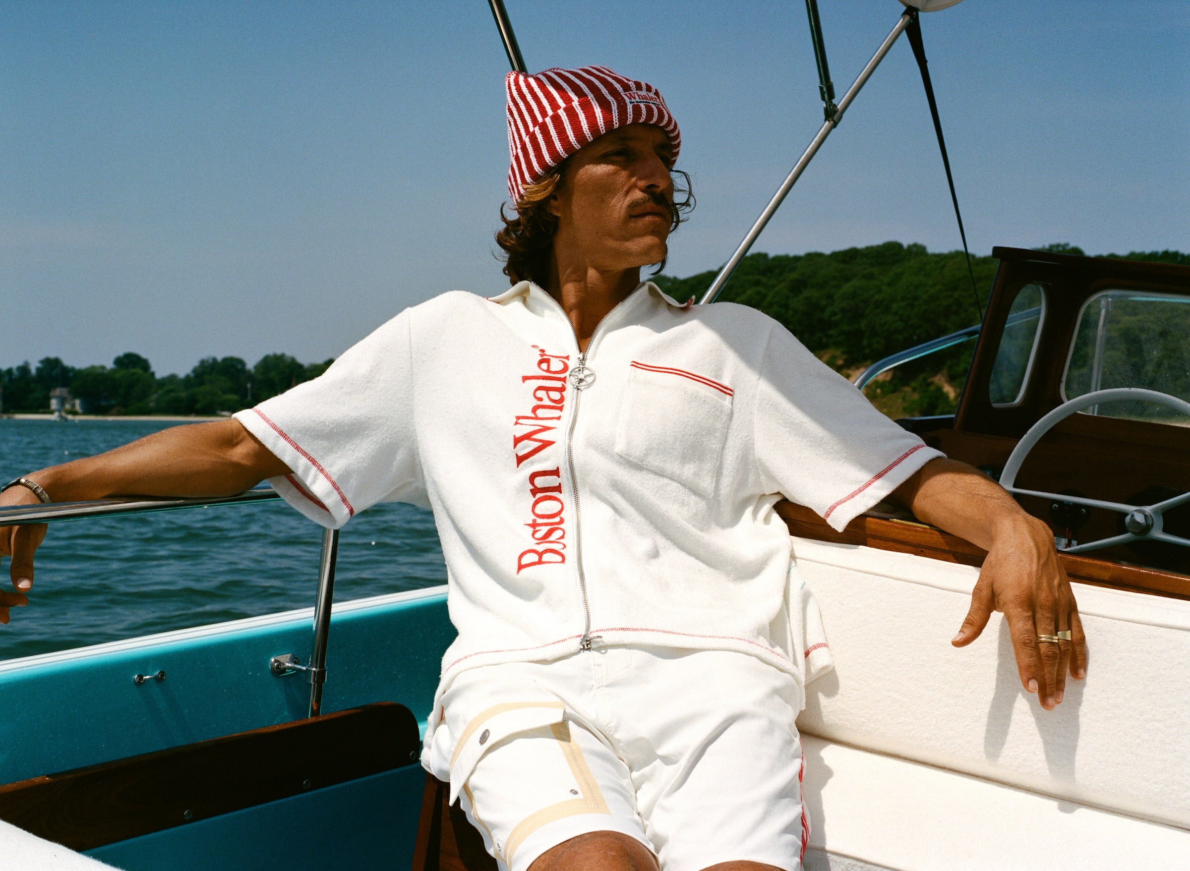 Man wearing white short-sleeve terry cloth zip-up shirt with red vertical 'Boston Whaler' text and red contrast stitching, while sitting on a boat. He also wears a red-and-white striped beanie with a 'Whaler' logo across the front.