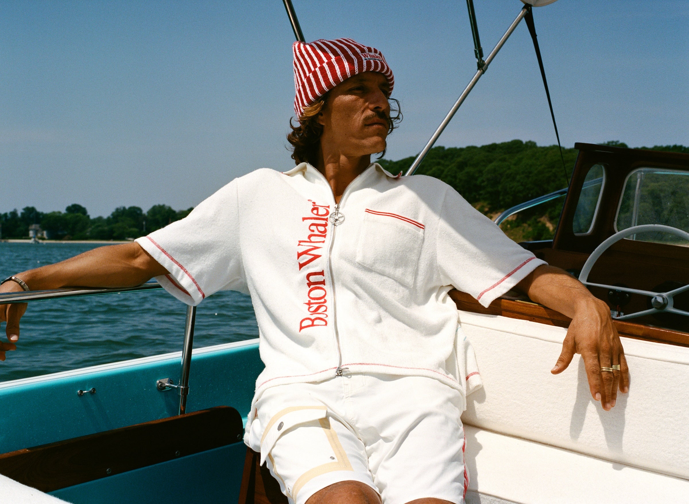 Man wearing white short-sleeve terry cloth zip-up shirt with red vertical 'Boston Whaler' text and red contrast stitching, while sitting on a boat. He also wears a red-and-white striped beanie with a 'Whaler' logo across the front.