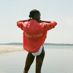 Back view of a woman standing in shallow water, wearing a red long-sleeve shirt with white embroidered text reading 'Boston Whaler.' The shirt is worn over a white swimsuit, and she is adjusting the collar with both hands.