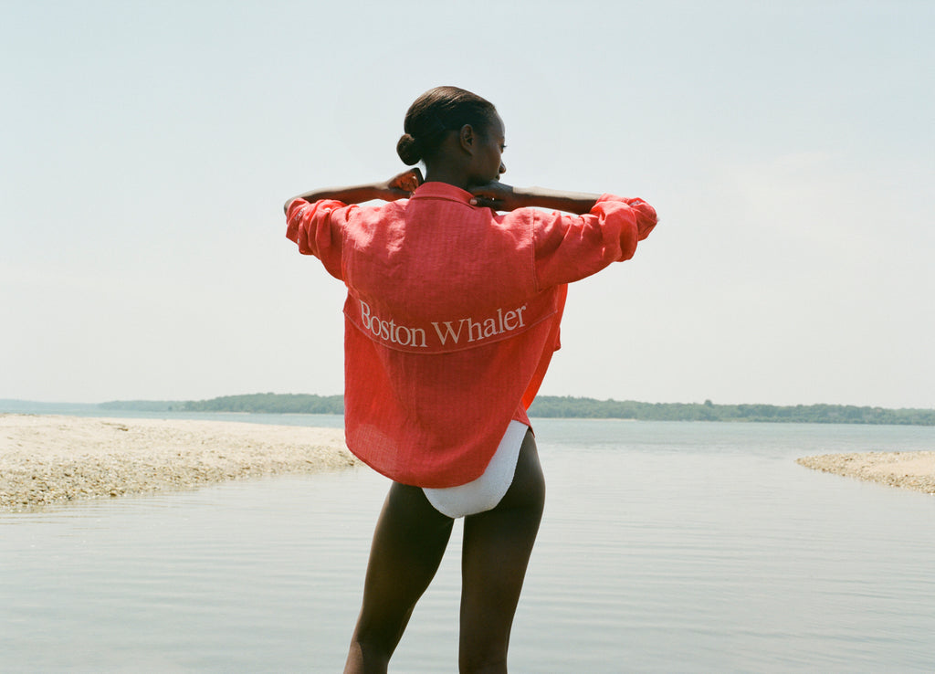Back view of a woman standing in shallow water, wearing a red long-sleeve shirt with white embroidered text reading 'Boston Whaler.' The shirt is worn over a white swimsuit, and she is adjusting the collar with both hands.