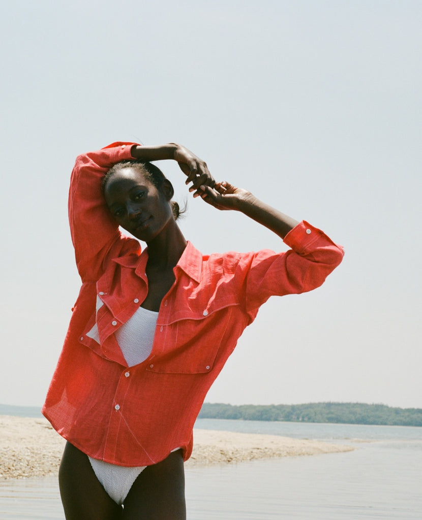 Woman standing on the beach, wearing a red button-up shirt over a white one-piece swimsuit. 