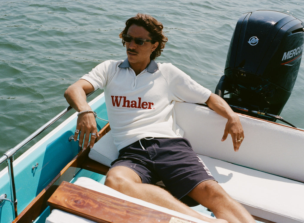 Man lounging on a boat, wearing a white 'Whaler' polo shirt and black shorts with 'Whaler' text on the side, and a pair of sunglasses. 
