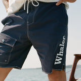 Close-up of man on boat wearing navy board shorts with white contrast stitching, and 'Whaler' text visible down the man's left leg. 