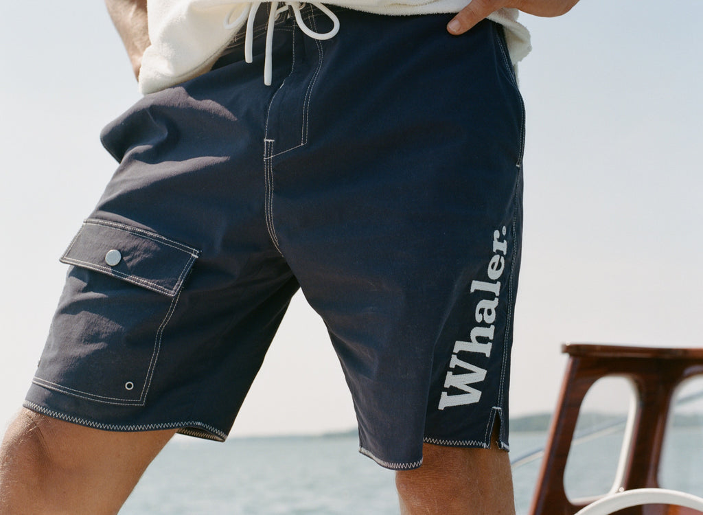 Close-up of man on boat wearing navy board shorts with white contrast stitching, and 'Whaler' text visible down the man's left leg. 