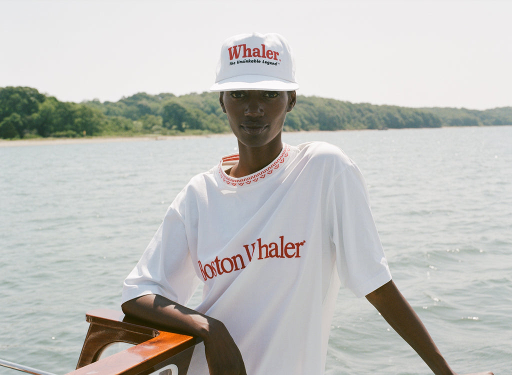 Woman on a boat wearing a white five-panel cap with red embroidered text reading 'Whaler' and smaller text reading 'The Unsinkable Legend'. A small red 'Tombolo' logo is visible on the side of the hat. She is also wearing a matching white 'Boston Whaler' shirt with red stitching at the collar.