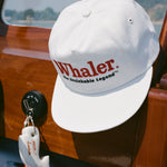 White baseball cap with red embroidered text reading 'Whaler' and black text below reading 'The Unsinkable Legend,' hanging inside a wooden boat. Below the hat, a whale-shaped white key float with 'Tombolo' branding is attached to a red Mercury engine kill switch.