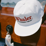 White baseball cap with red embroidered text reading 'Whaler' and black text below reading 'The Unsinkable Legend,' hanging inside a wooden boat. Below the hat, a whale-shaped white key float with 'Tombolo' branding is attached to a red Mercury engine kill switch.