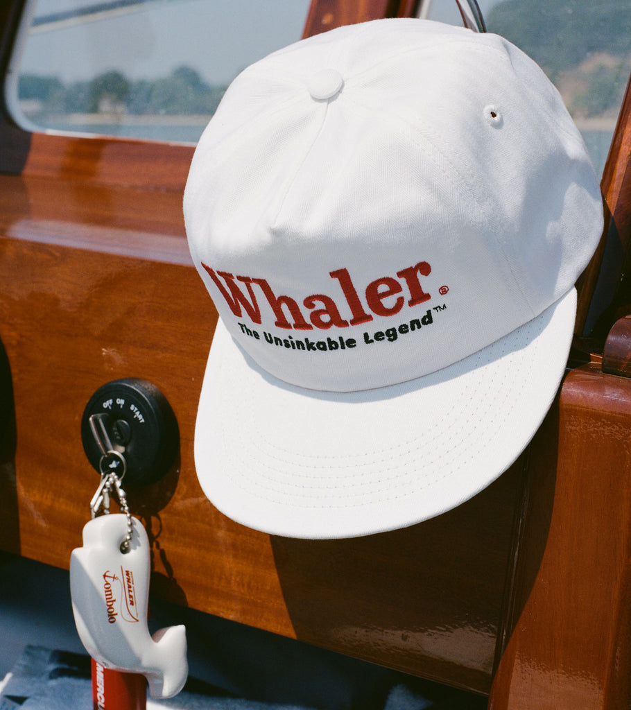 White baseball cap with red embroidered text reading 'Whaler' and black text below reading 'The Unsinkable Legend,' hanging inside a wooden boat. Below the hat, a whale-shaped white key float with 'Tombolo' branding is attached to a red Mercury engine kill switch.