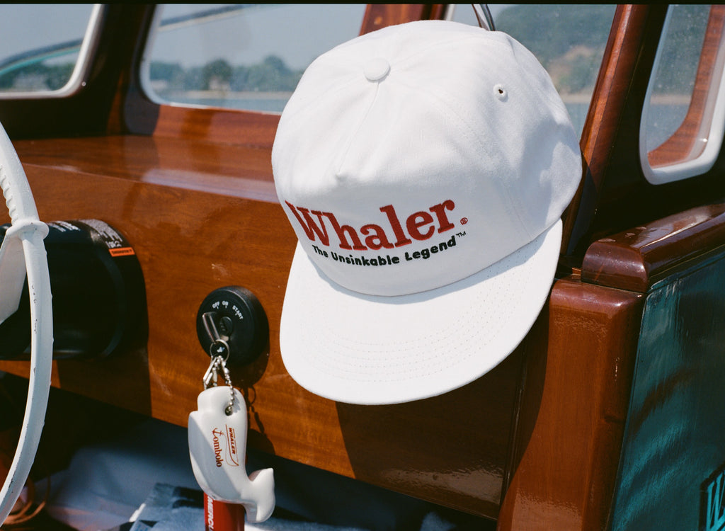White baseball cap with red embroidered text reading 'Whaler' and black text below reading 'The Unsinkable Legend,' hanging inside a wooden boat. Below the hat, a whale-shaped white key float with 'Tombolo' branding is attached to a red Mercury engine kill switch.